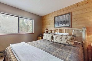 Een slaapkamer met een houten hoofdeinde, een groot bed met een ruitbeddengoed, twee kussens met hertenmotieven, een nachtkastje en een ingelijst beeld aan de muur.