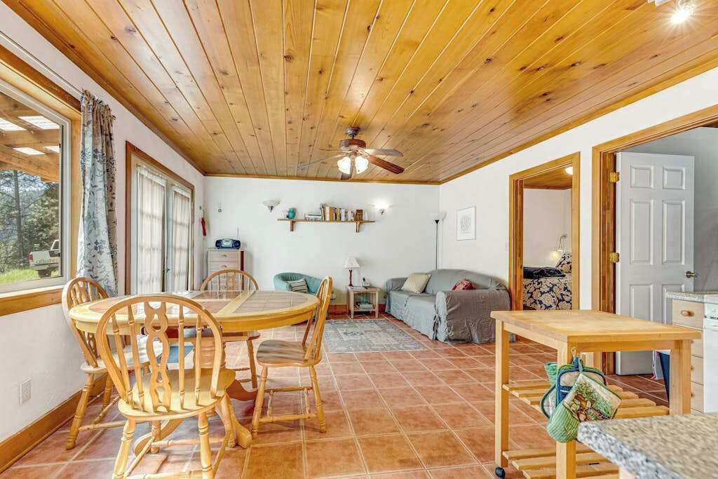 Una habitació acollidora amb sostre de fusta, taula de dinar amb caires, un sofà, una petita taula auxiliar i una cuina visible a través d'una porta oberta.