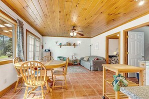Ein gemütliches Zimmer mit Holzbalkendecke, einem Esstisch mit Stühlen, einer Couch, einem kleinen Beistelltisch und einem durch eine geöffnete Tür einsehbarer Küchenbereich.