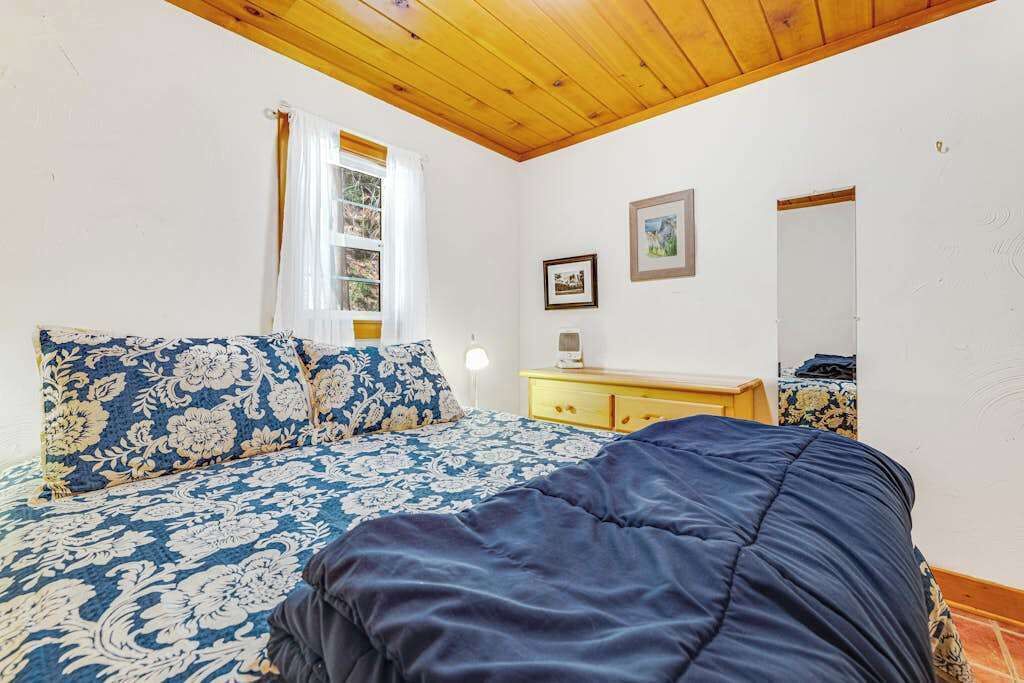 A bedroom with a bed, a wooden ceiling, a window with curtains, a bedside table, and a wall-mounted mirror.