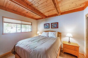 Ein Schlafzimmer mit Holzbalkendecke, einem Bett mit Kopfteil, einem Nachttisch, zwei Wandleuchten und einem Fenster mit Jalousien.