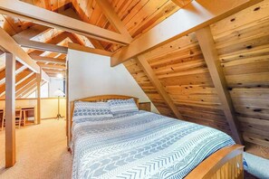 A bedroom with a sloped ceiling, a bed with patterned bedding, and a desk with a lamp. - Pine Paradise - Big Trees Villag (Camp Connell)