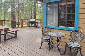 Eine Holzterrasse mit Tisch, zwei Stühlen, einem Grill und einem großen Fenster.