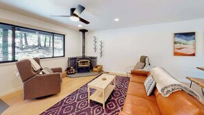 A modern living room with a fireplace, a sofa, a recliner, and a large window offering a view of trees. - Piney Point - Meadowmont (Arnold)