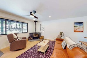 Ein modernes Wohnzimmer mit Kamin, einer Couch, einem Relaxsessel und einem großen Fenster mit Blick auf Bäume.