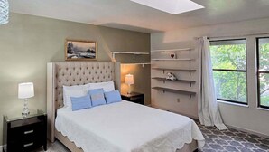 A bedroom with a tufted headboard, a bed with white and blue bedding, a bedside lamp, a nightstand, a shelf with decorative items, and a window with a view of greenery. - Pleasant Pines Retreat - Pine Mountain Lake (Groveland)