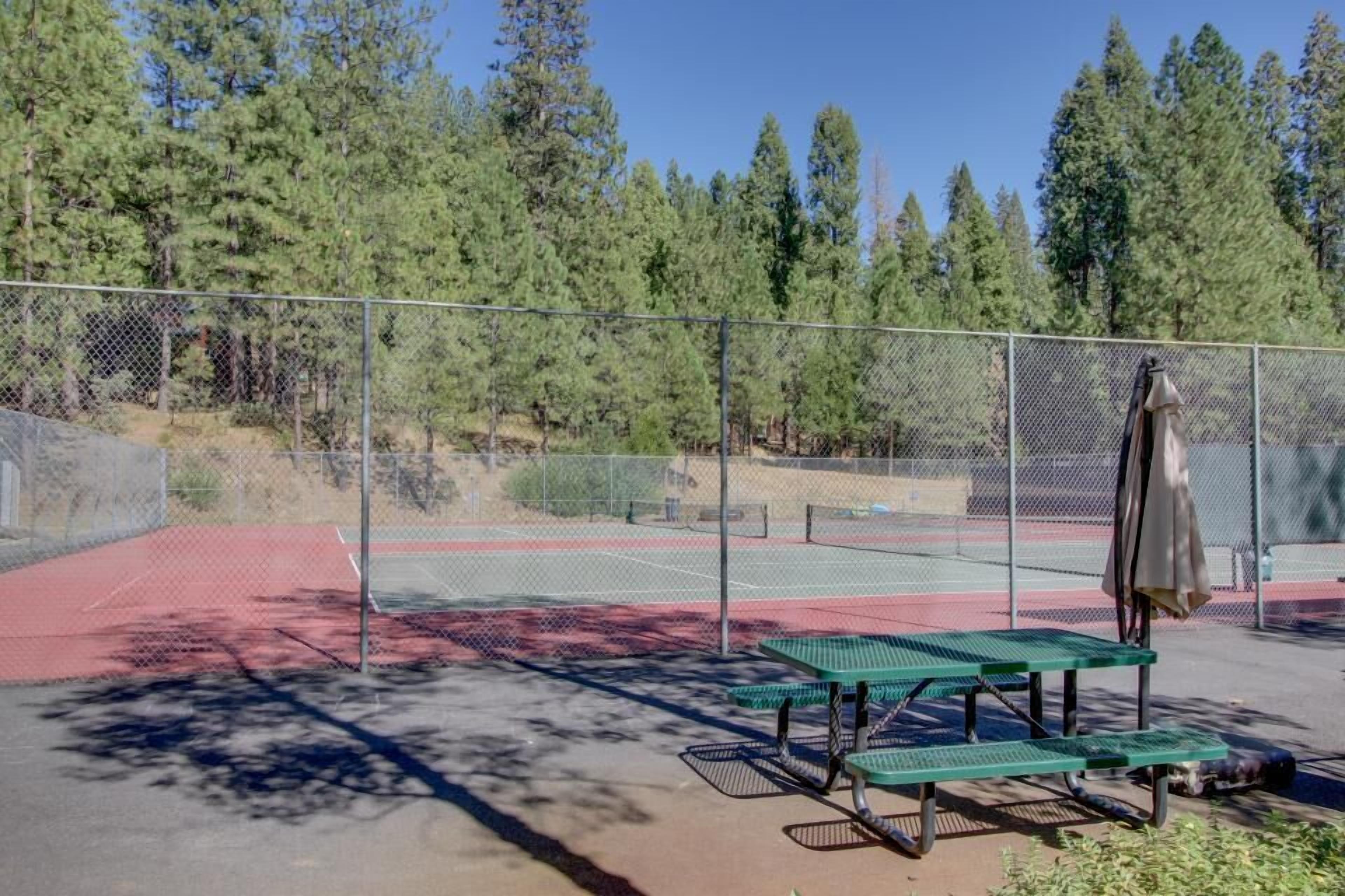 Cancha de tenis al aire libre con una mesa de picnic y un banco, rodeada por una cerca de malla y árboles.