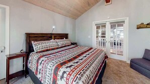 A bedroom with a large bed, wooden headboard, a nightstand, a desk, a chair, and a door with glass panels. - Windwood Inn - Ockenden Ranch (Shaver Lake)