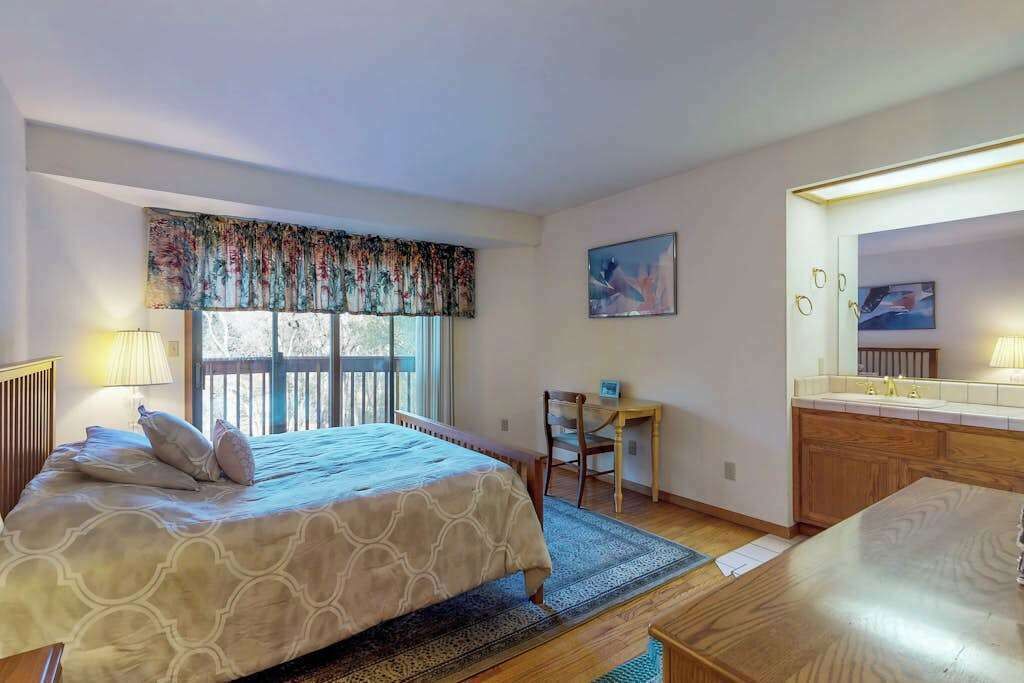 A bedroom with a bed, a desk, a chair, a window with curtains, a painting, and a bathroom with a mirror and sink.