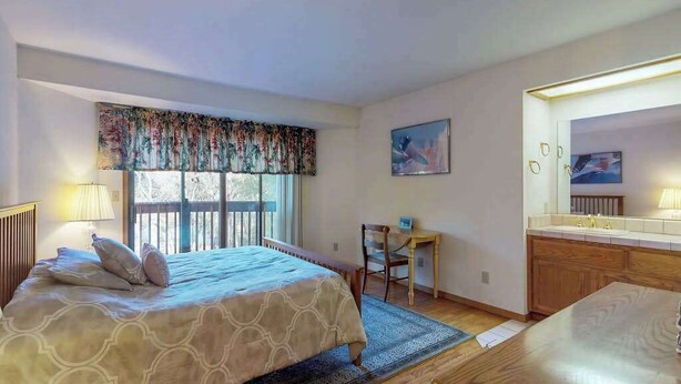 A bedroom with a bed, a desk, a chair, a window with curtains, a painting, and a bathroom with a mirror and sink. - Lake Lodge Retreat - Pine Mountain Lake (Groveland)