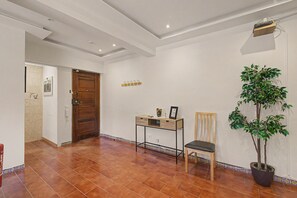 Un couloir avec une porte en bois, une petite table avec une chaise, une plante en pot et une étagère fixée au mur.