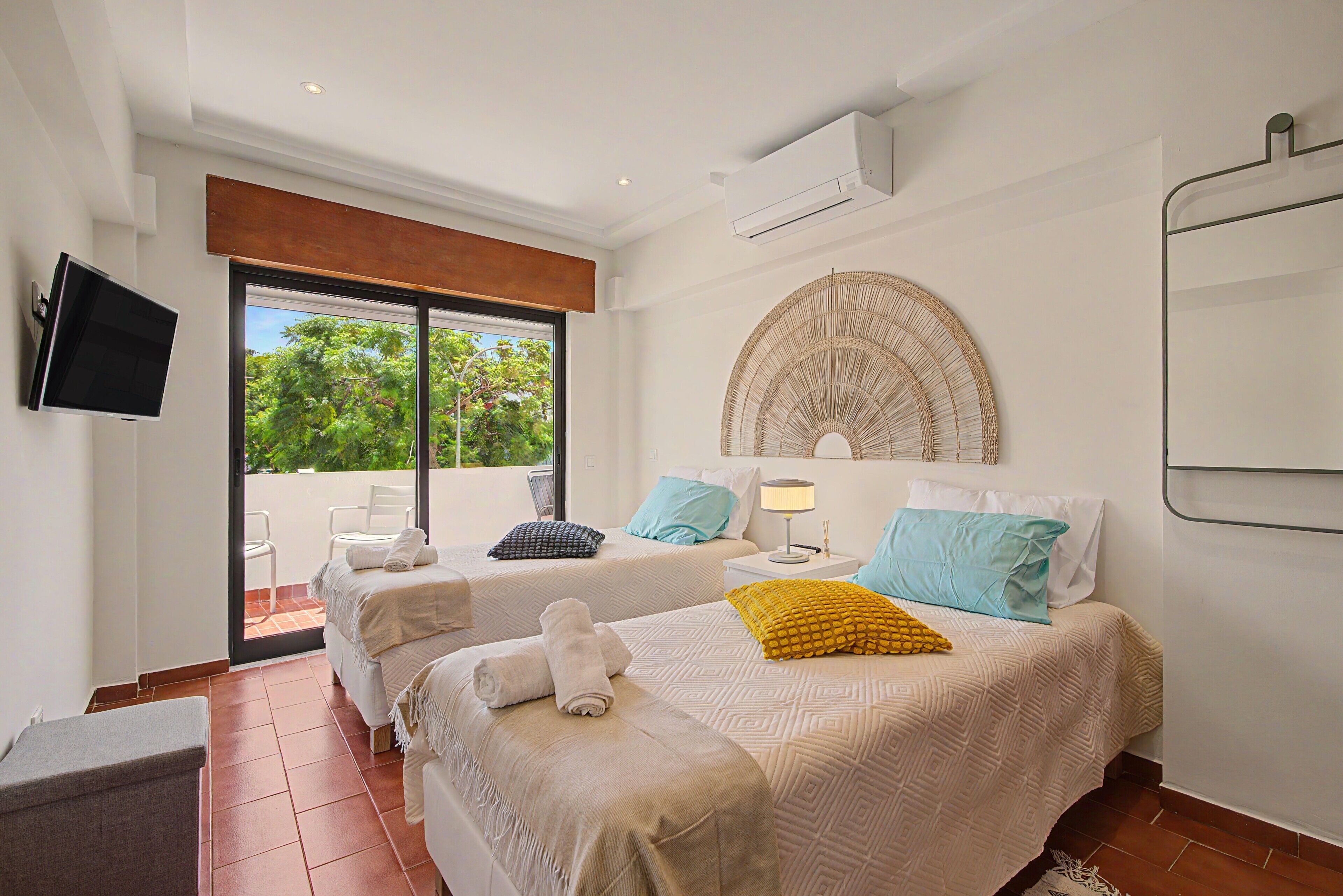 A hotel room with two beds, a TV, a window with a view of greenery, and a ceiling fan.