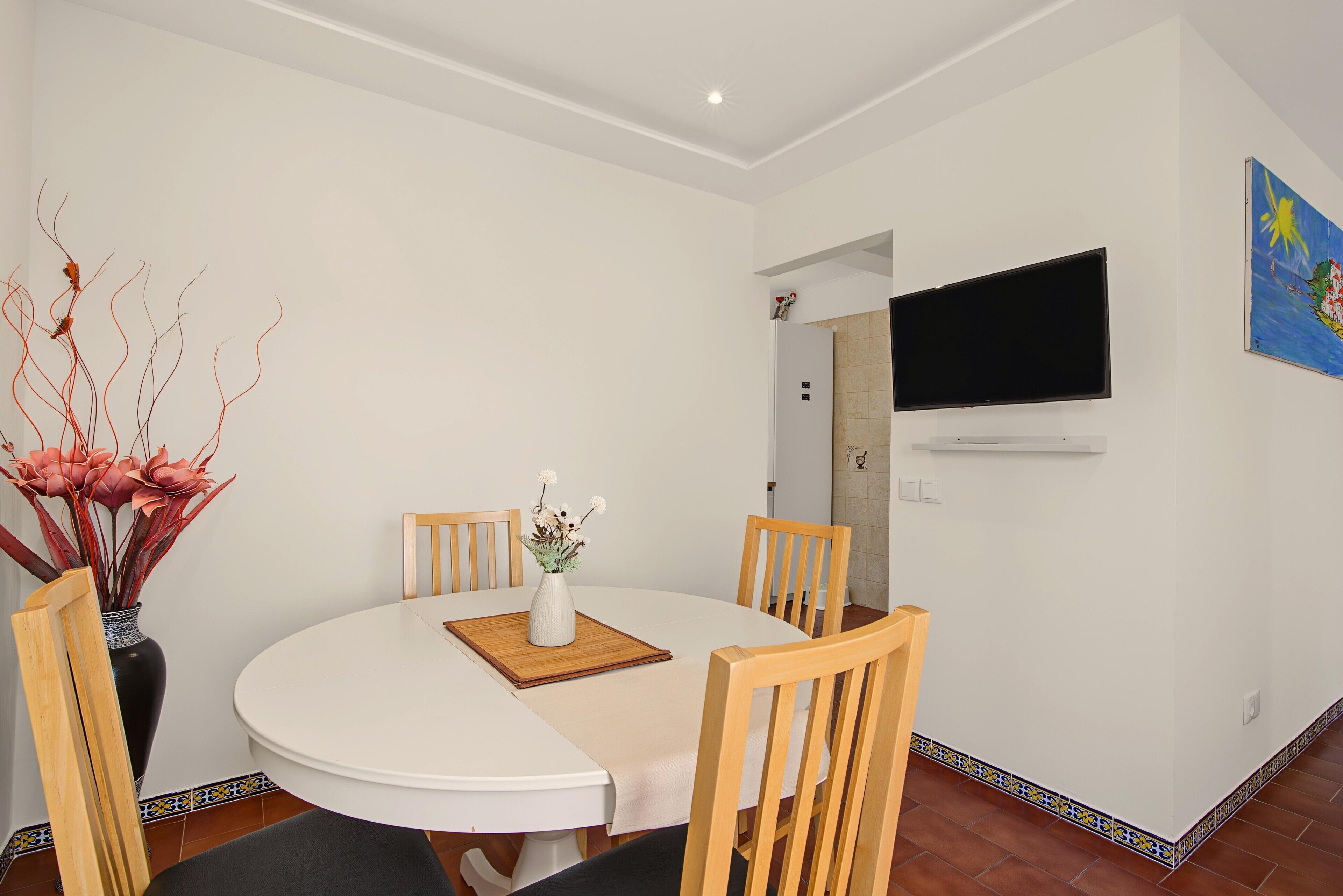Een eetgedeelte met een ronde tafel, vier houten stoelen, een witte vaas met bloemen en een flatscreen aan de muur.