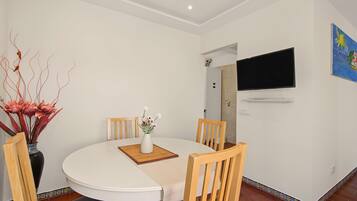 Een eetgedeelte met een ronde tafel, vier houten stoelen, een witte vaas met bloemen en een flatscreen aan de muur.