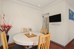 Uma sala de jantar com uma mesa redonda, quatro cadeiras de madeira, um vaso branco com flores e uma televisão de tela plana na parede.