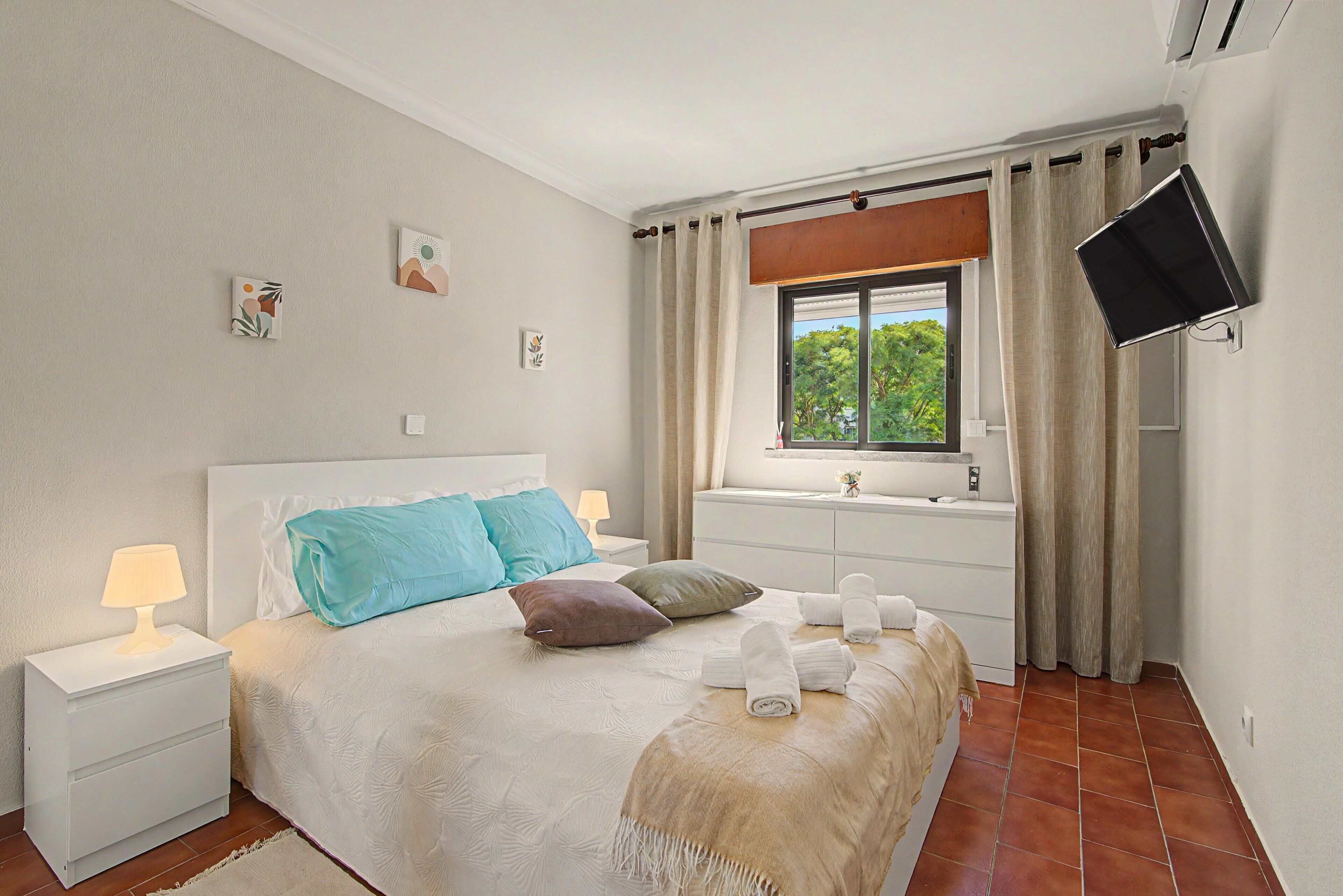 A bedroom with a bed, bedside tables, a window with curtains, a flat-screen TV, and a small bedside lamp.