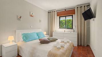 A bedroom with a bed, bedside tables, a window with curtains, a flat-screen TV, and a small bedside lamp.