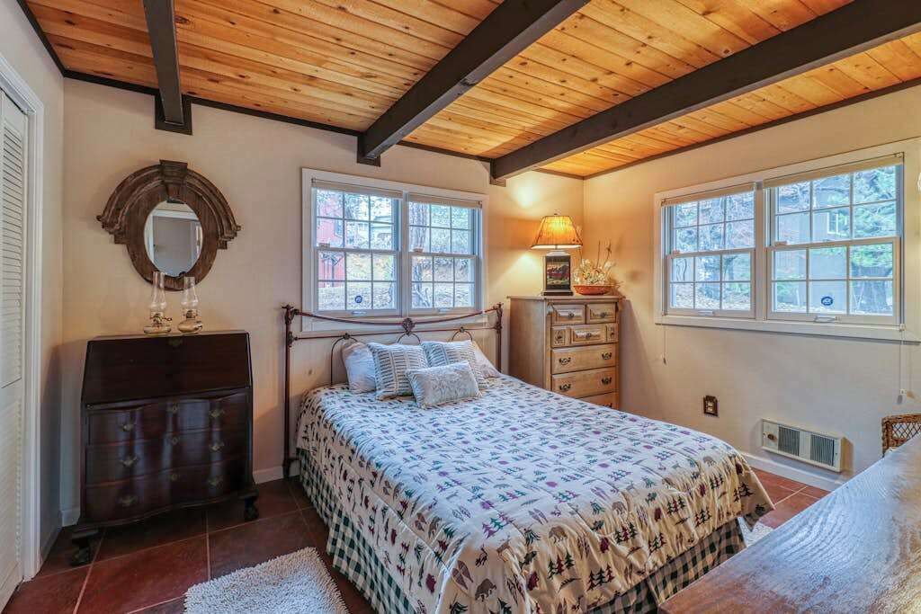Une chambre avec un plafond en bois, un lit recouvert d’une couvre-lit à motifs, une commode, un miroir et une fenêtre donnant sur des arbres.