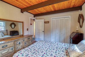 Ein Schlafzimmer mit Holzbalkendecke, einem Bett mit gemusterten Textilien, einem Schminktisch, einem Spiegel und einem Kleiderschrank.