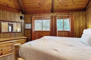 A wooden cabin bedroom with a bed, a dresser, a mirror, and a window with curtains. - Rustic Retreat - Pine Mountain Lake (Groveland)