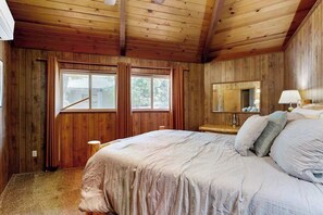 Une chambre à coucher avec un plafond en bois, un grand lit avec des oreillers, une table de chevet, un miroir et deux fenêtres avec des rideaux.