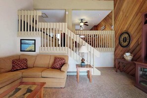 Ein Wohnzimmer mit einem beigefarbenen Ecksofa, einem kleinen Holztisch, einer Lampe und einer Treppe mit Holzbalken.