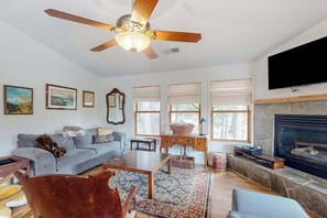 A living room with a ceiling fan, a fireplace, a television mounted on the wall, and a sofa with a dog. - Under The Stars - Shaver Lake (Shaver Lake)