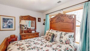 Ein Schlafzimmer mit Holzkopfende, Blumenbettwäsche, einem Nachttisch mit Lampe und einem fenster mit Vorhängen.