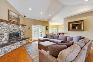 A living room with a stone fireplace, a sectional sofa, and a dining area in the background. - Mule Deer Cabin - Pine Mountain Lake (Groveland)