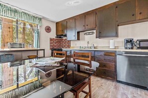 A kitchen with a brick wall, wooden cabinets, a glass table, and a dishwasher. - Big Trees, Little Cabin - Meadowmont (Arnold)