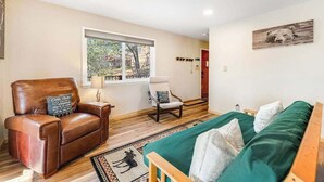 Ein Zimmer mit Ledersessel, grünem Sofa, Fenster mit Blick auf Bäume und Wandbild.
