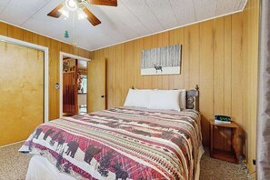 Un dormitorio con pared de paneles de madera, una cama con colcha estampada, un reloj sobre una mesita de noche y un ventilador de techo.
