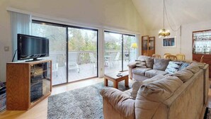 Un salón amplio con una televisión grande sobre un soporte de madera, un sofá color beige, una mesa de centro y un balcón con muebles de exterior.
