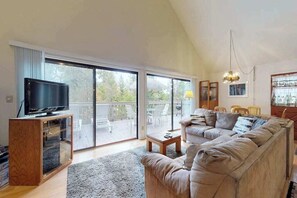 Un salon spacieux avec une grande télévision posée sur un meuble en bois, un canapé beige, une table basse et un balcon aménagé avec du mobilier d’extérieur.