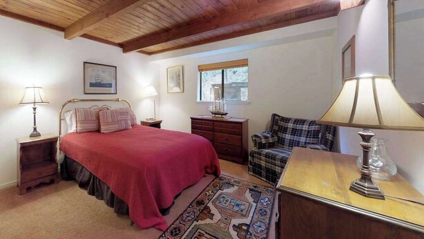 Un dormitorio con techo de madera, cama con colcha roja, cómoda, silla y lámpara.