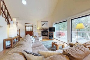 A spacious living room with a large sofa, a fireplace, a television, and a balcony with outdoor seating. - Forest Chalet - Pine Mountain Lake (Groveland)