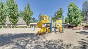 Un parque infantil con un tobogán espiral amarillo y estructuras de escalada azules, rodeado de árboles y un edificio al fondo.