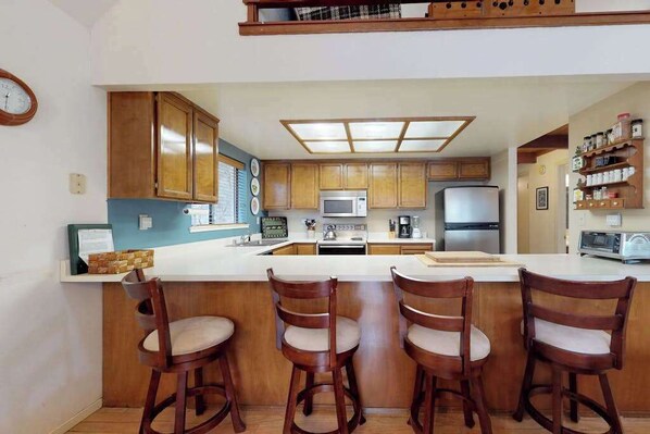 A kitchen with wooden cabinets, a central island with bar stools, a microwave, a refrigerator, and a clock on the wall.