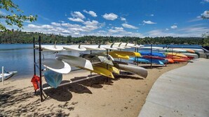 Una hilera de canoas y kayaks alineados en una playa arenosa junto a un lago tranquilo.