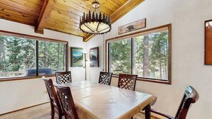 A dining area with a wooden table and chairs, large windows with a forest view, and a chandelier. - On The 9th - Blue Lake Springs (Arnold)