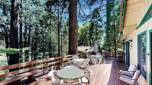 An outdoor deck with wooden flooring, surrounded by tall trees, featuring outdoor seating and a wooden railing. - On The 9th - Blue Lake Springs (Arnold)