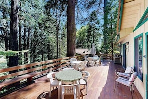 Eine Terrasse im Freien mit Holzboden, umgeben von hohen Bäumen, mit Sitzgelegenheiten und einem Holzgeländer.