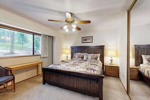 A bedroom with a large bed, a ceiling fan, bedside lamps, a bench, and a window with a view of trees.