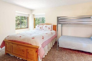 Quarto com cama de madeira, beliche e duas janelas com vista para o verde.