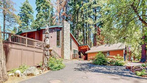A cabin with a stone chimney, wooden deck, and a paved driveway surrounded by trees. - Tranquil Pines Retreat - Ridgetop (Shaver Lake)