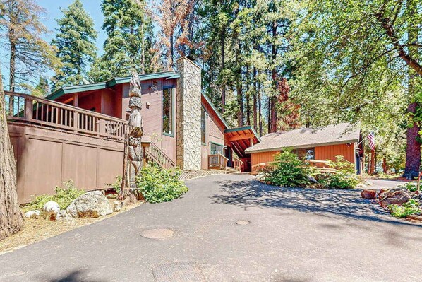 A cabin with a stone chimney, wooden deck, and a paved driveway surrounded by trees. - Tranquil Pines Retreat - Ridgetop (Shaver Lake)