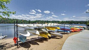 Una hilera de canoas y kayaks alineados en una playa arenosa junto a un lago tranquilo.