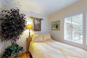 Ein Schlafzimmer mit einem Bett, einem Nachttisch, einer Lampe, einer großen Kübelpflanze und einem Fenster mit Jalousien.