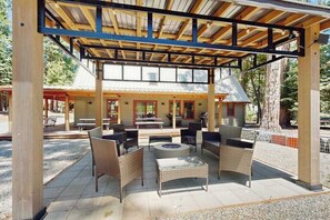 An outdoor patio area with wicker furniture and a fire pit, surrounded by a covered structure with a metal roof. - Sparrow Cabin - East Village (Shaver Lake)