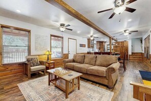 A spacious living room with a large sofa, a coffee table, and a fireplace. There are ceiling fans, wooden beams, and a kitchen area in the background. - Sparrow Cabin - East Village (Shaver Lake)